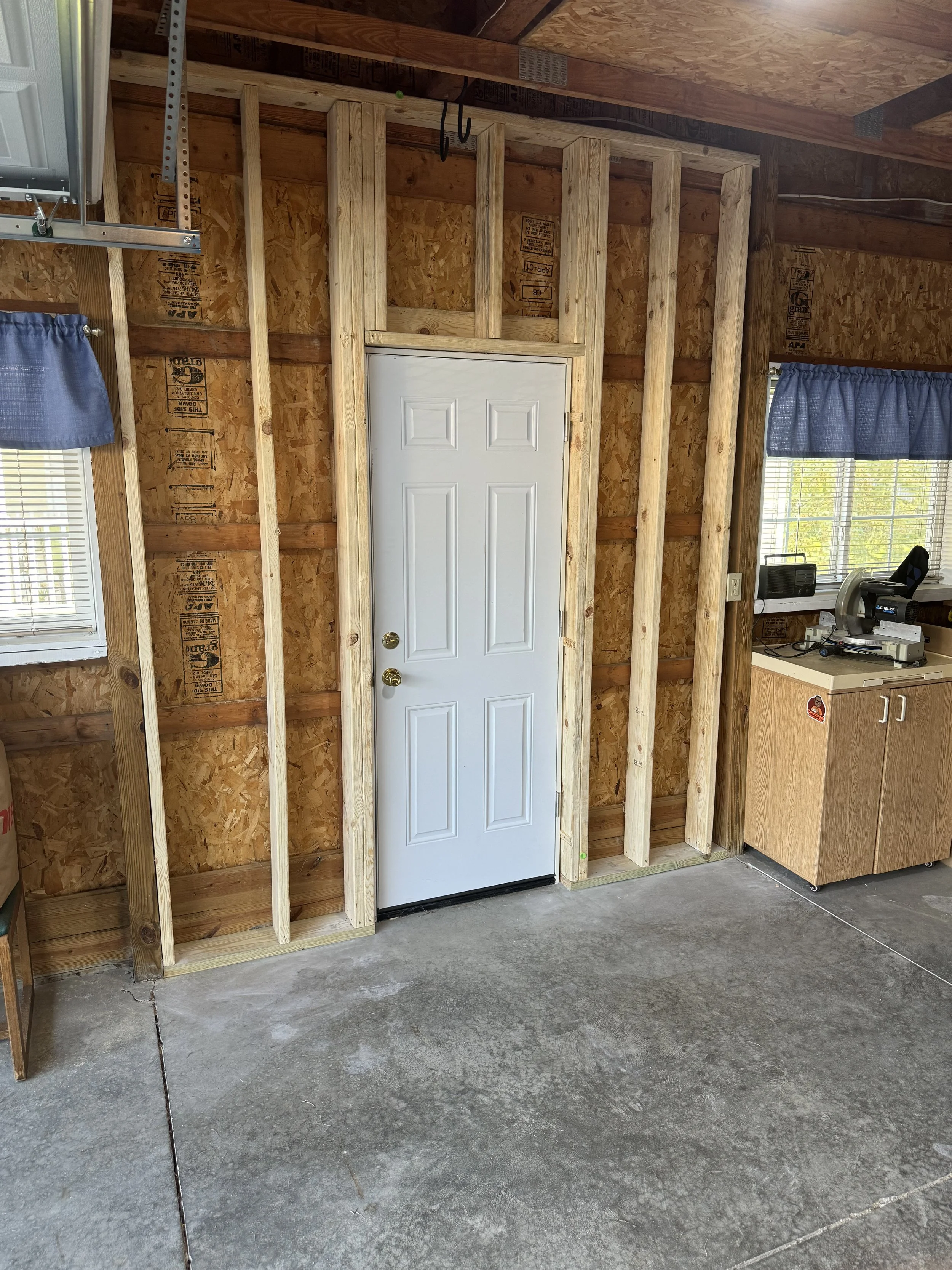 Garage workshop with unfinished wall framing and a white exterior door, concrete floor, two windows with blue curtains, and a workbench with a miter saw.