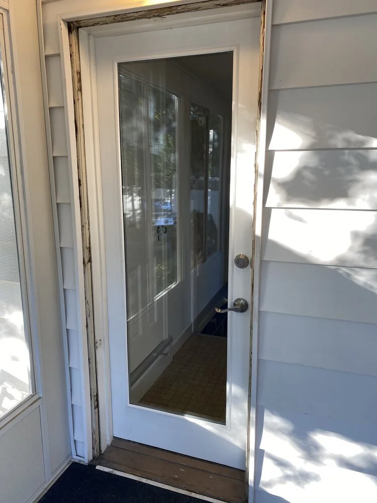 An exterior view of a door with a glass panel, with damaged or missing trim around the door frame, on a house with white siding.
