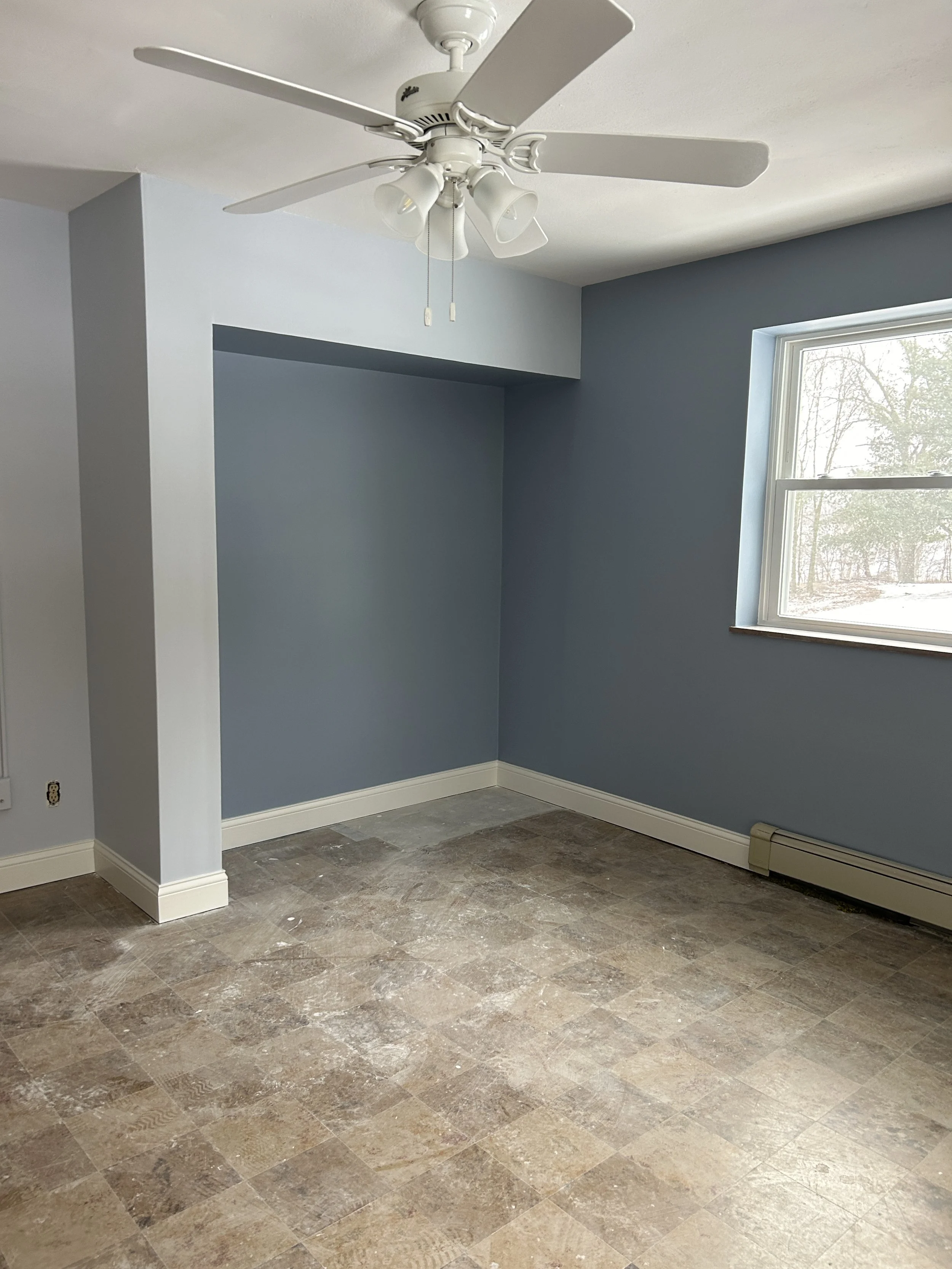 Empty room with blue walls, a window showing outside trees, a ceiling fan with attached light fixtures, and a partially carpeted floor.