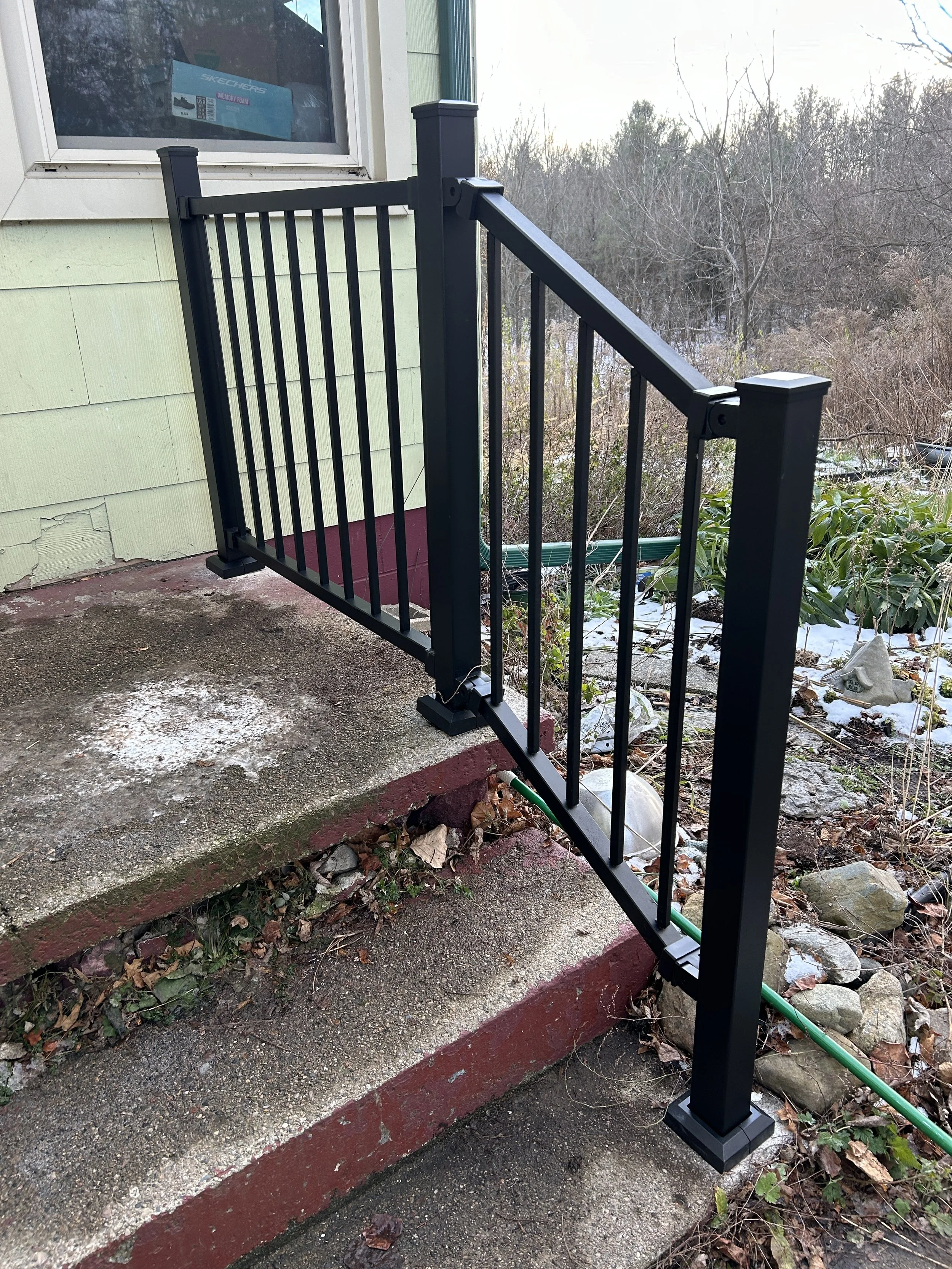 New black metal safety railing installed on outdoor concrete stairs adjacent to a house with light green siding and a window.