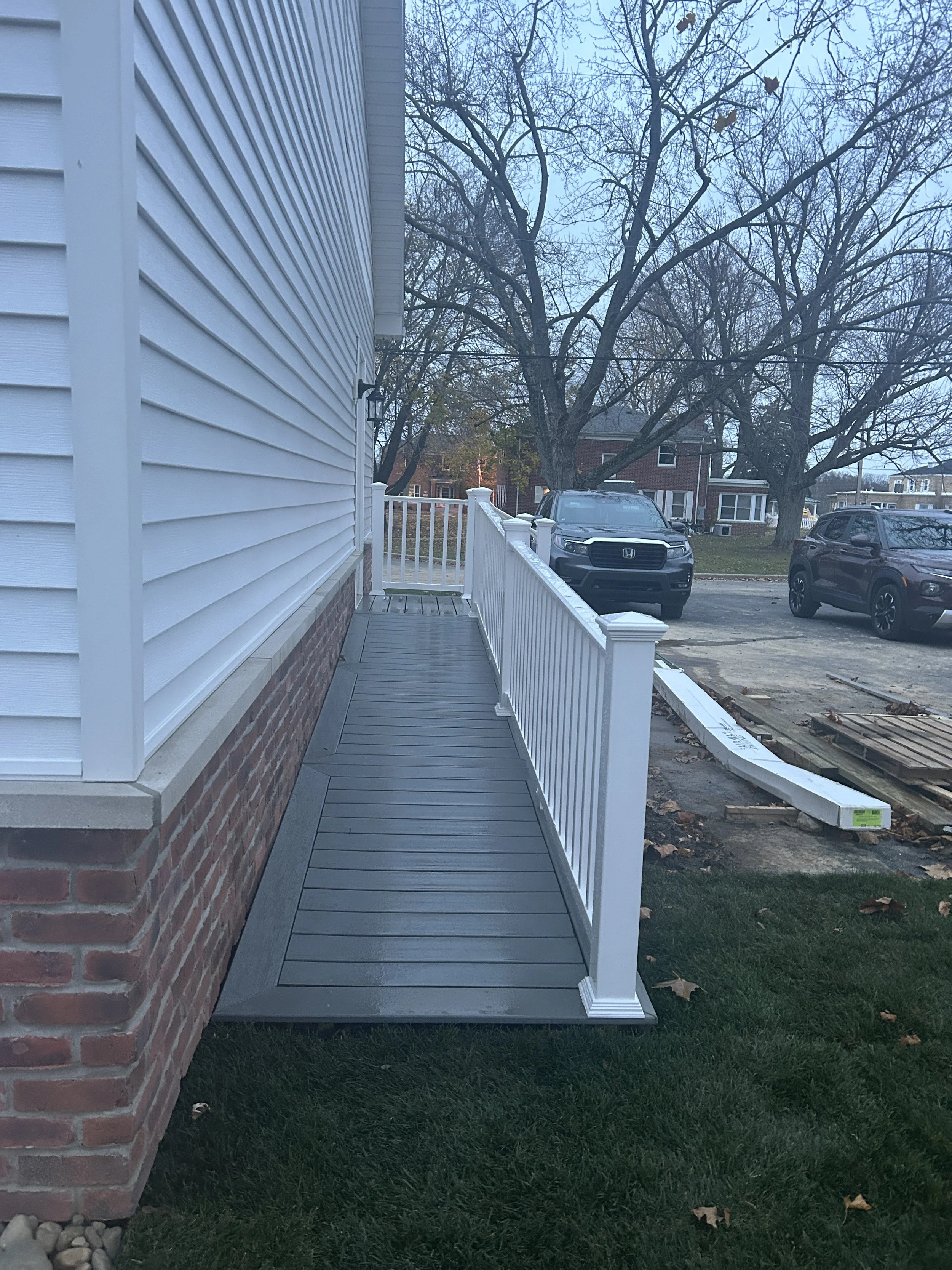 Residential porch with new grey composite deck, white railing, and siding on the house, with cars parked on the street and fallen leaves around.