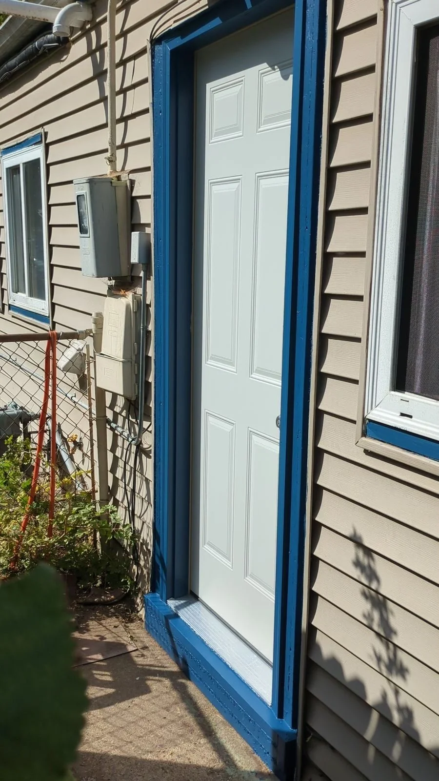 Exterior view of a house door with a white door and a blue painted frame, next to a window with a similar blue and silver frame, beige siding, an electrical meter and boxes, and a small fenced garden with plants and shadows cast by nearby objects.