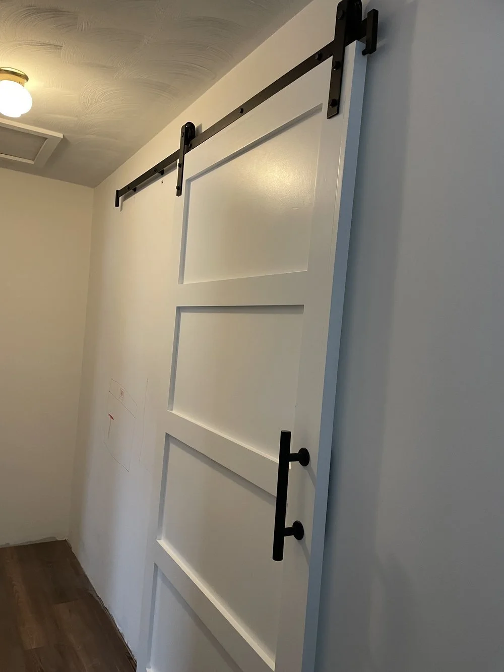 A white sliding barn door with three rectangular panels, black hardware, and a black handle, installed on a wall near the ceiling of a room with a wooden floor and a ceiling light.
