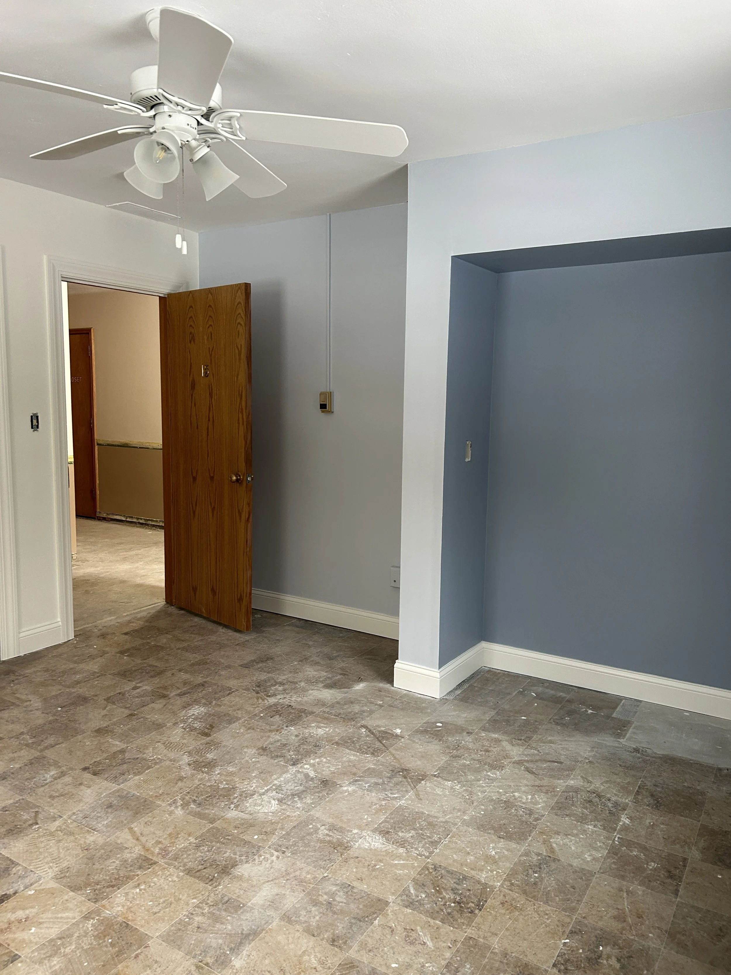 Empty room with tan tile flooring, light blue and white walls, a ceiling fan, and a partially open wooden door leading to another room with similar flooring.