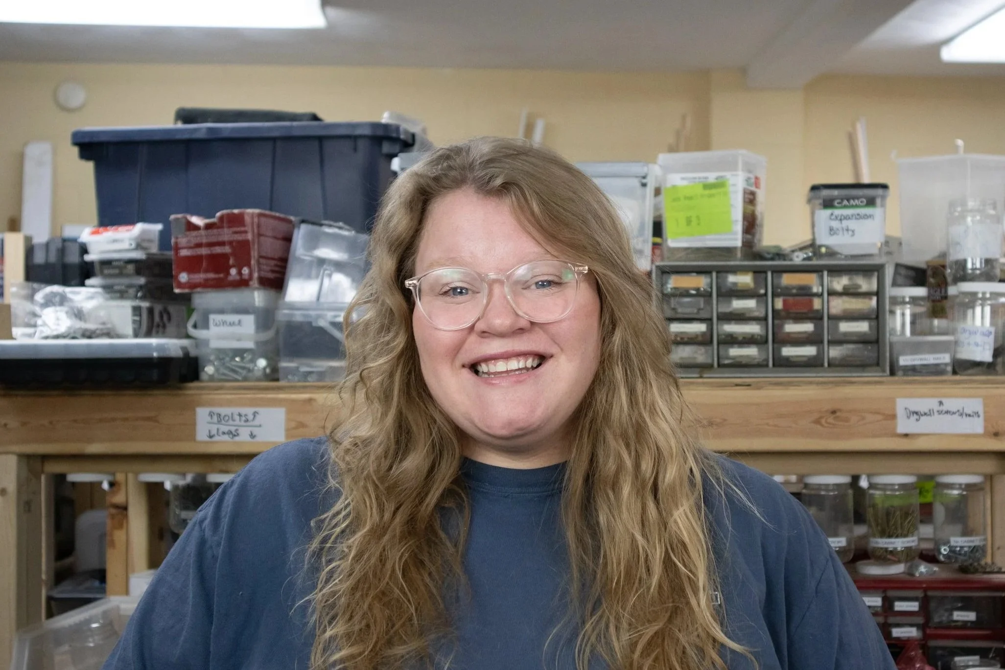 A woman with long, wavy red hair, wearing glasses and a dark blue shirt, smiling in a workshop or storage room with labeled shelves and plastic containers in the background.