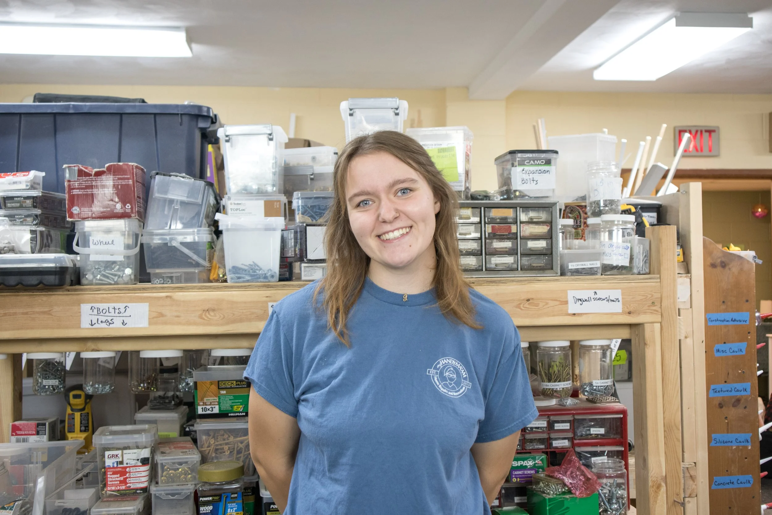 A young woman with long, wavy brown hair and a blue T-shirt smiling inside a hardware store with shelves of screws, bolts, and construction supplies.