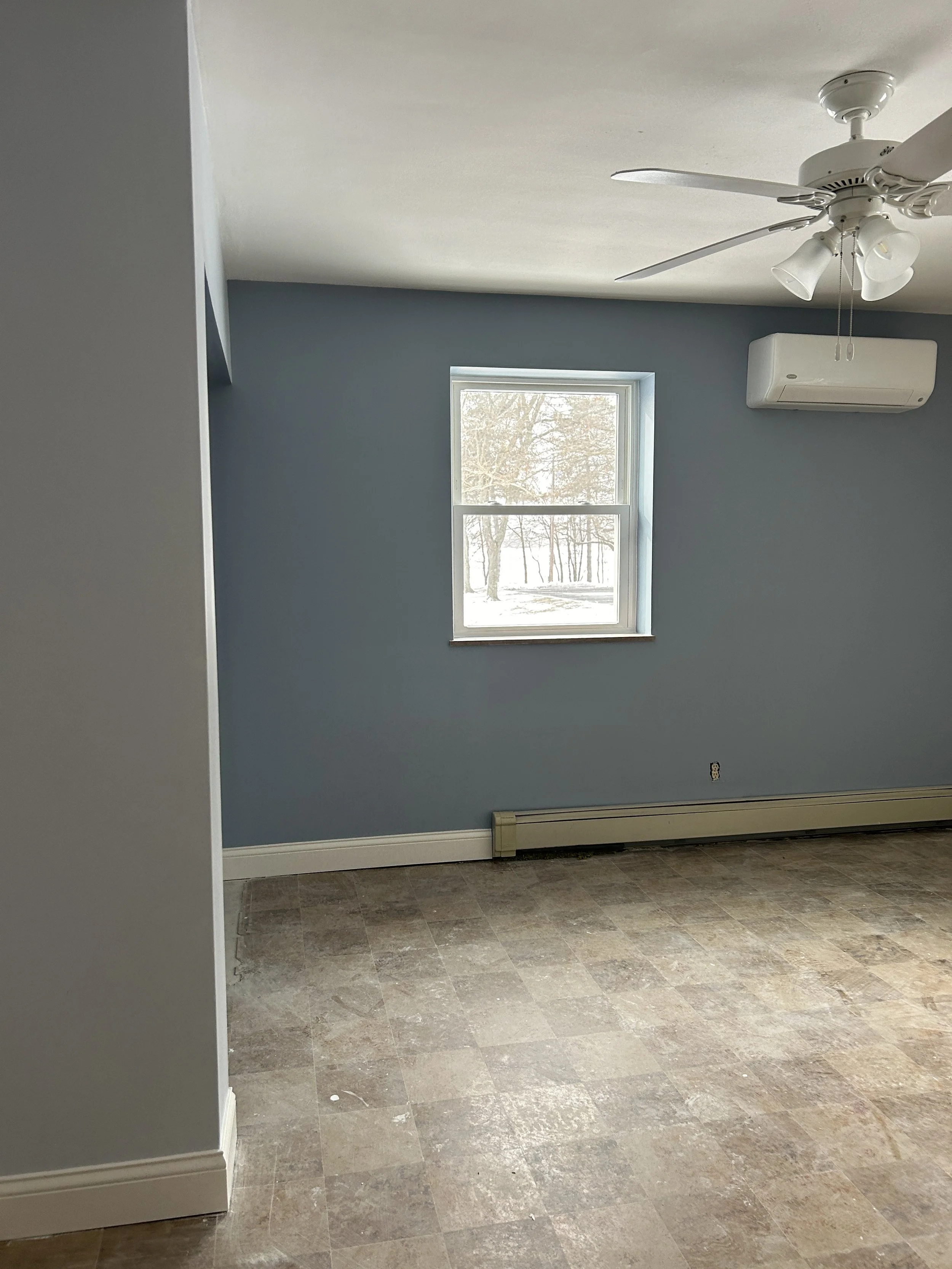 Empty room with light blue wall, window showing snowy outdoors, ceiling fan with lights, wall-mounted air conditioner, and beige tile floor.