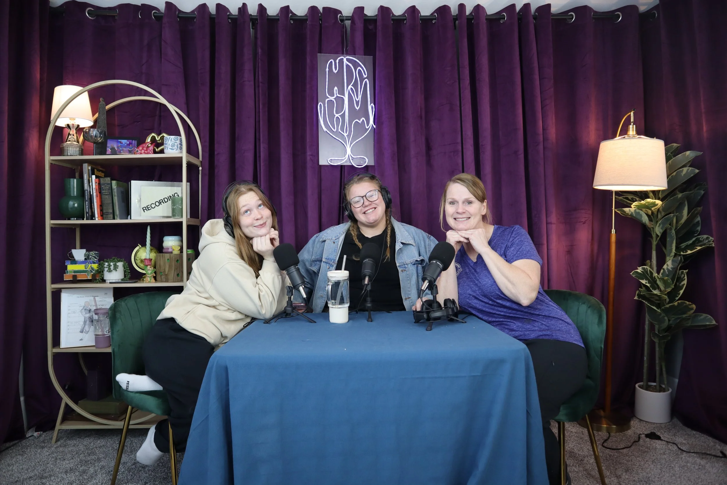 Three women sitting at a table with microphones, recording a podcast in a room with purple curtains, a bookshelf, and warm lighting.