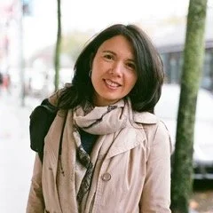 A woman smiling outdoors, wearing a coat and scarf, with trees and a blurred background.