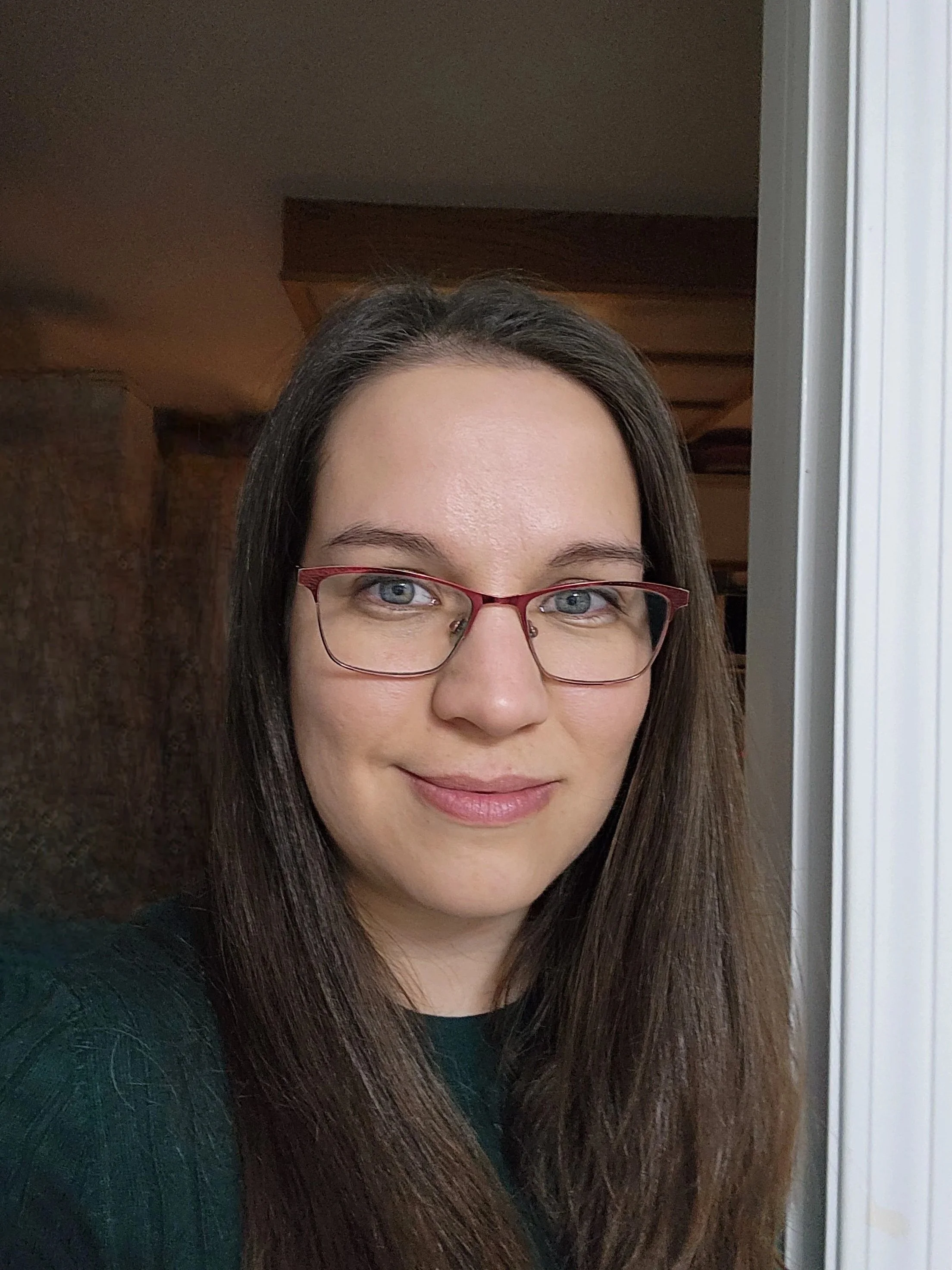 A woman with glasses and shoulder-length brown hair standing in front of a teal curtain, wearing a brown leather jacket, a gray scarf, and a red plaid shirt.
