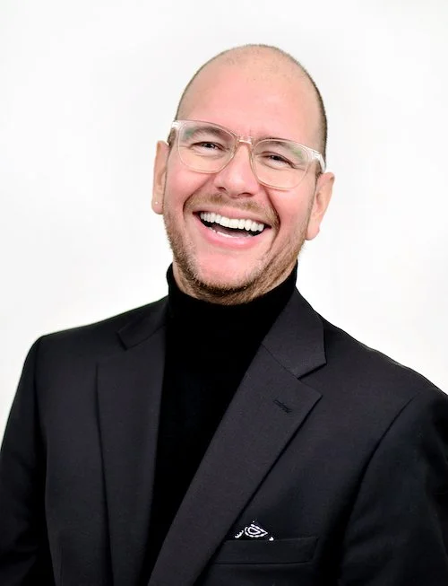 A person with a human face dressed in a banana costume, waving with a green glove against a gray background.
