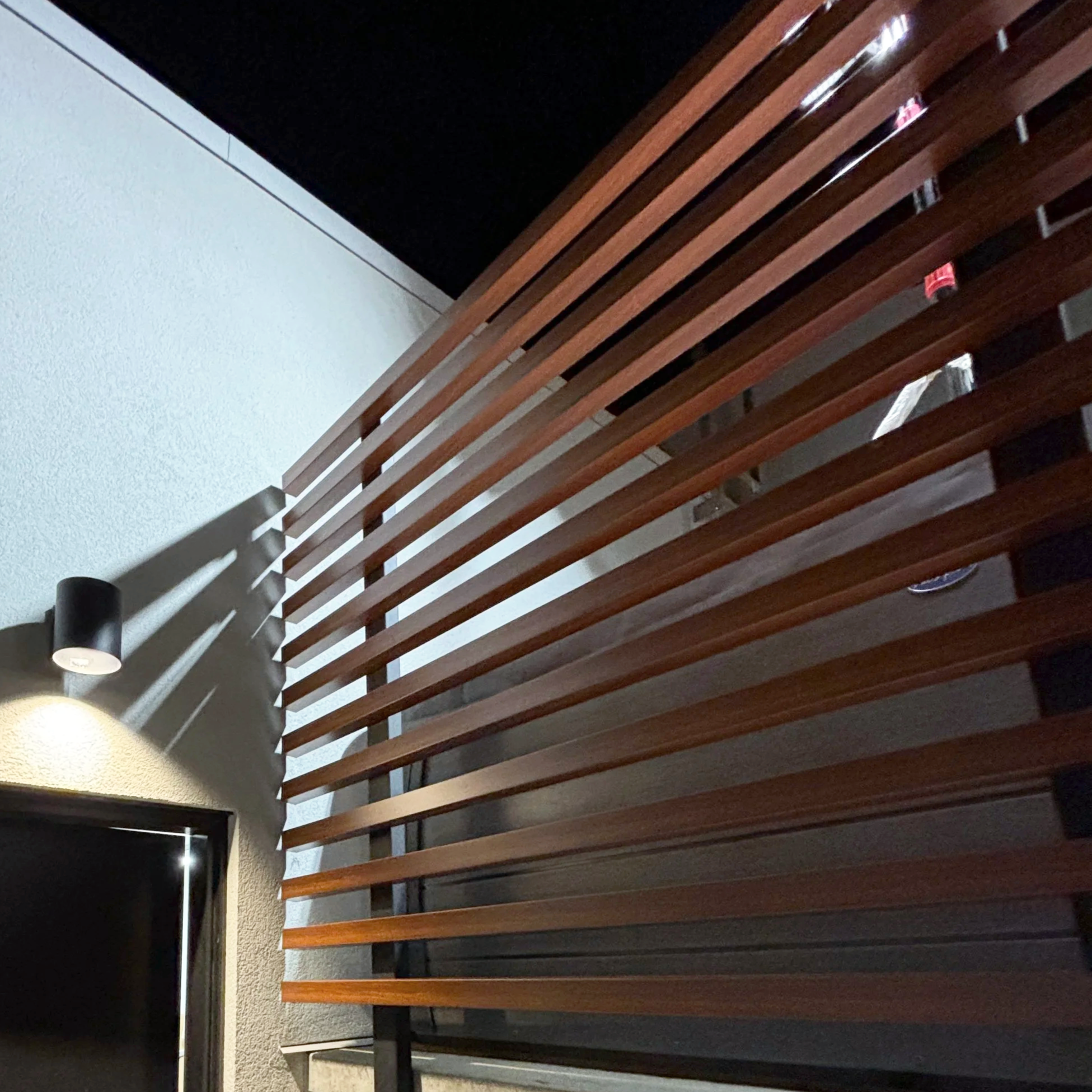 Close-up of wooden slats forming a privacy screen near a wall at night, with a wall-mounted light casting shadows.