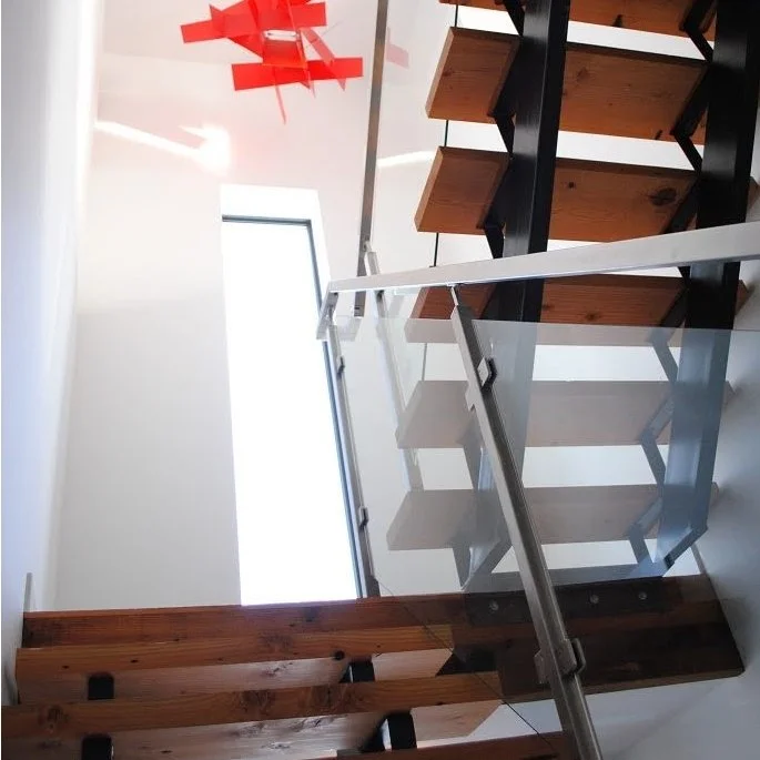 Looking up the staircase with wooden steps, metal railing, and glass panels, with a large window and a red ceiling light fixture.