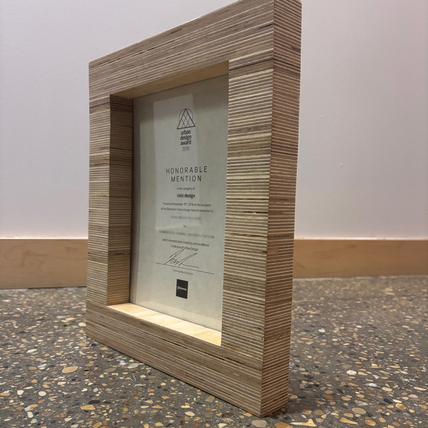 A laminated wooden frame award plaque, placed on a speckled floor against a wall, recognizing an honorable mention at the Edmonton Urban Design Awards.