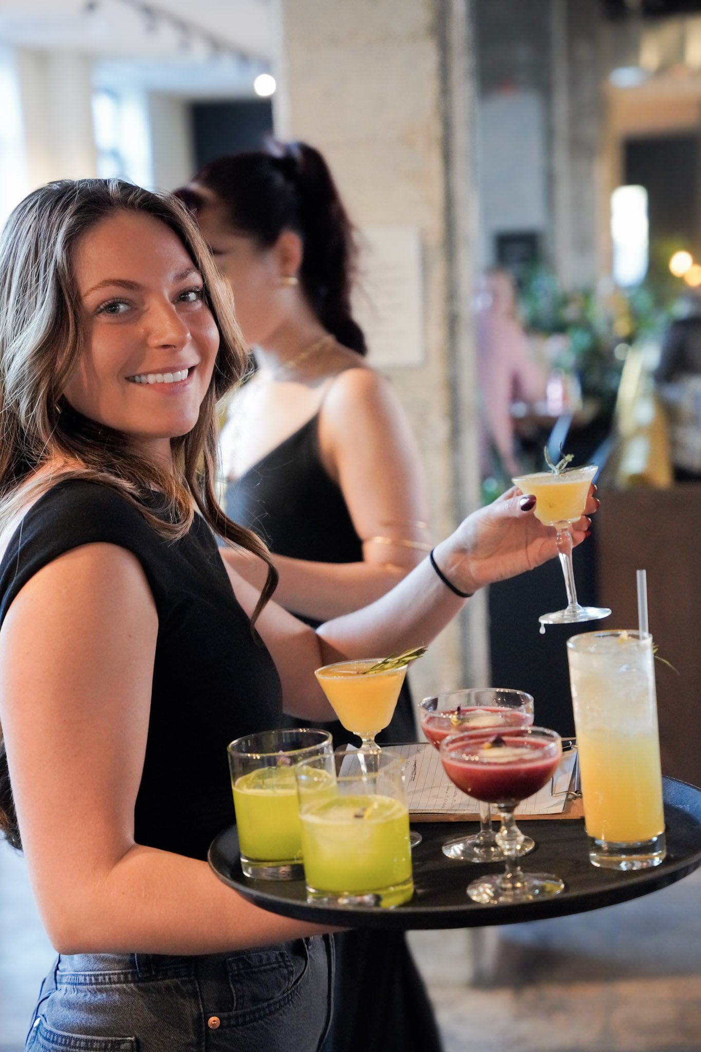 A smiling woman holding a tray of colorful cocktails at a bar or restaurant.