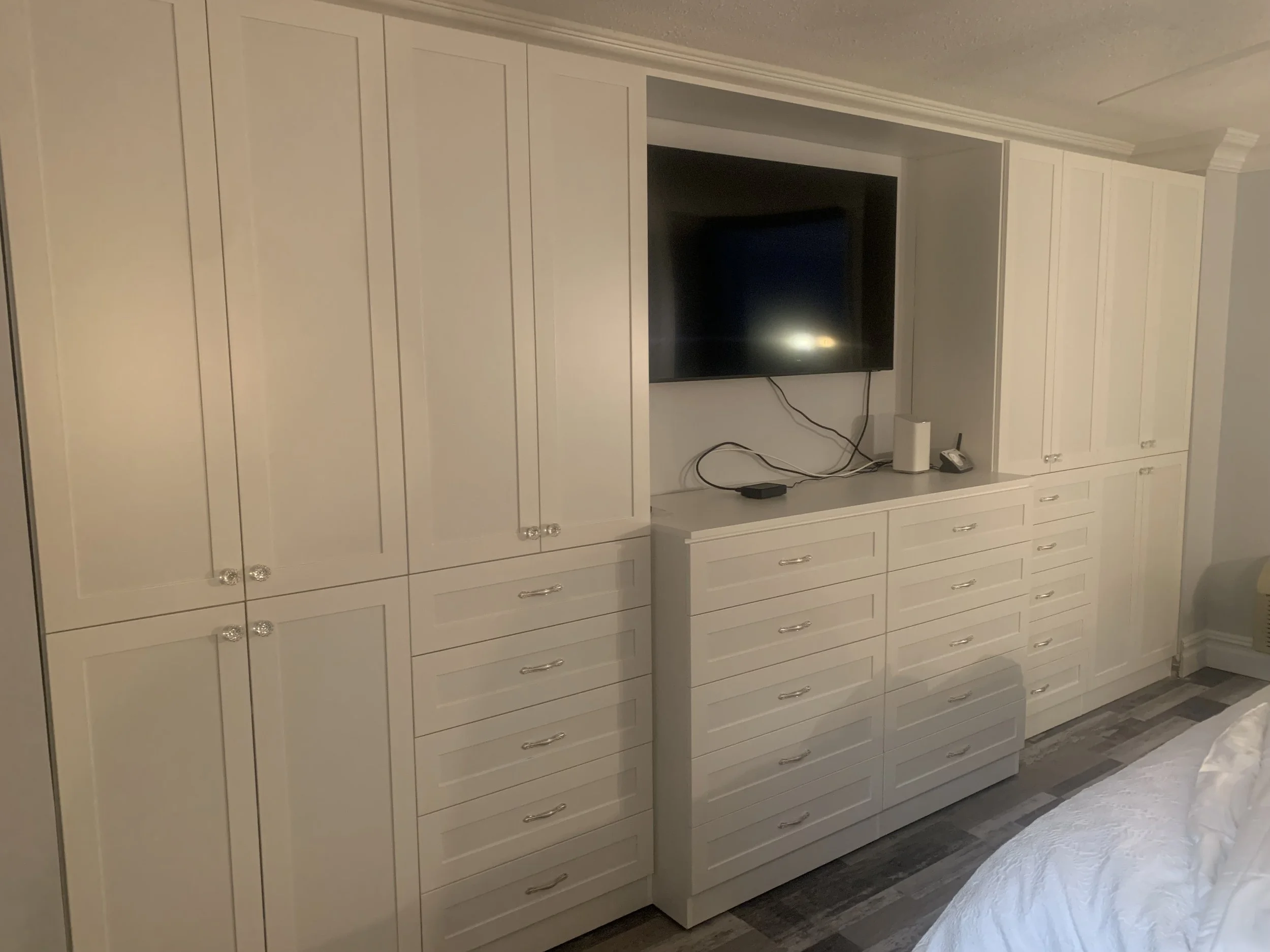 A large white built-in cabinet with multiple drawers and closed doors, occupying the entire wall, with a flat-screen TV mounted in the middle. Devices and cords are on the shelf below the TV.