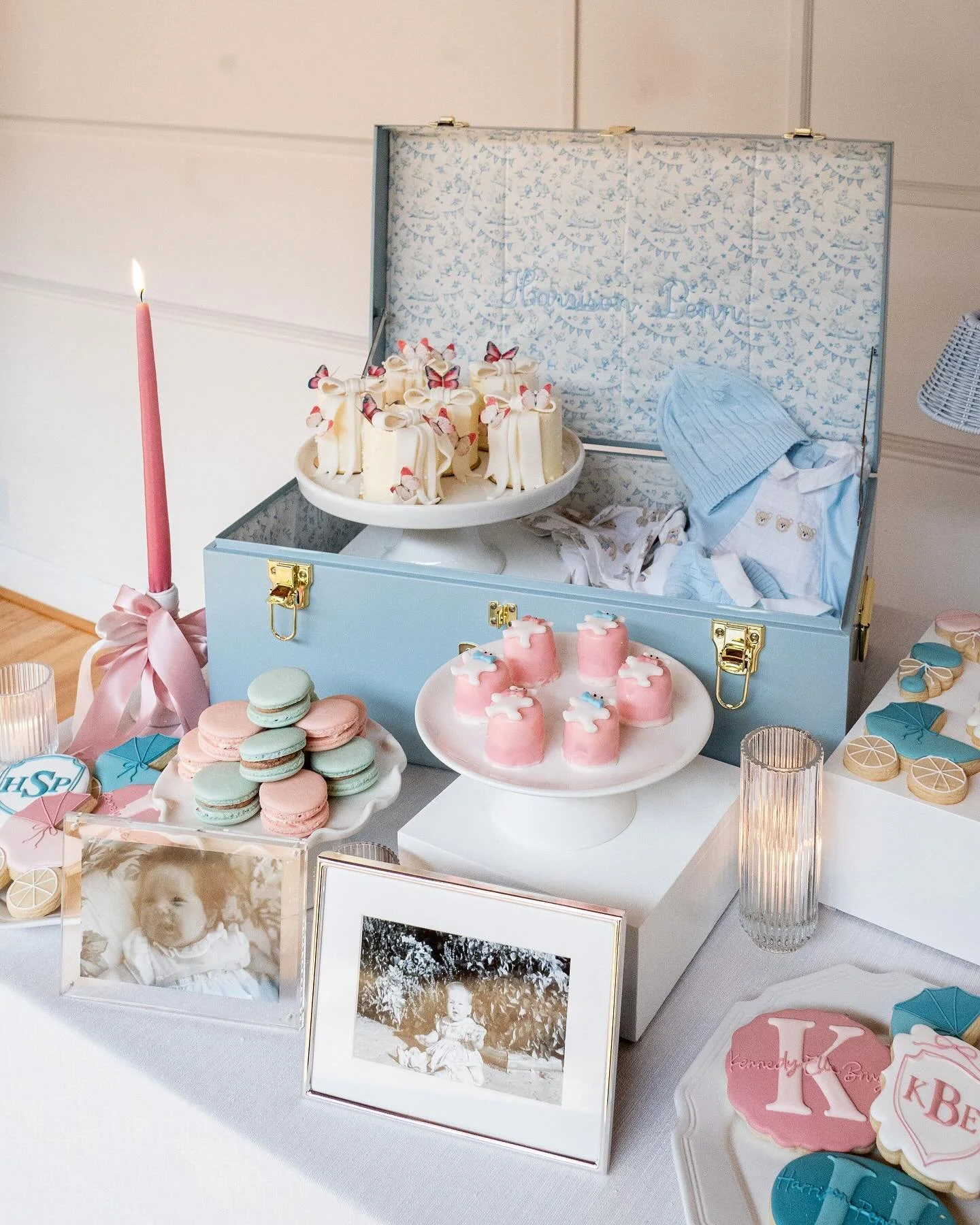 Table with pink, blue, and white baby-themed desserts and decorations, including framed black-and-white photos of a baby, a pastel blue box with baby clothing inside, and macarons in pastel colors.