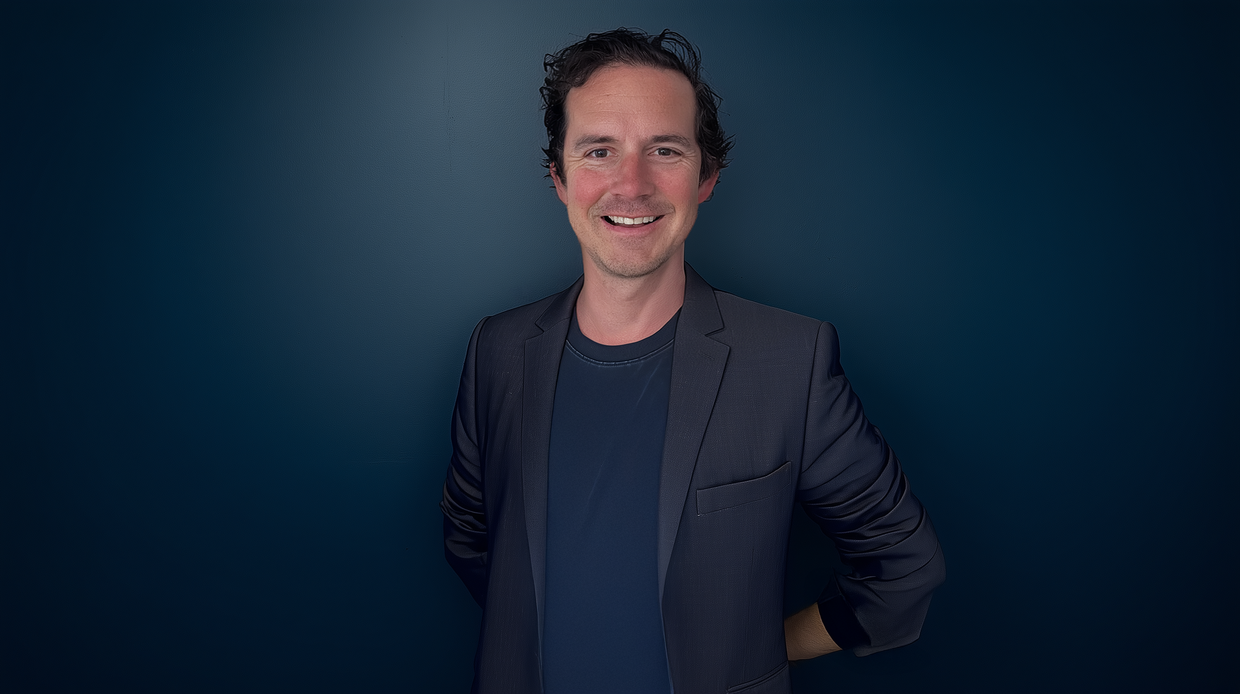 Smiling man with dark curly hair wearing a dark blazer over a dark shirt, standing against a dark blue background.