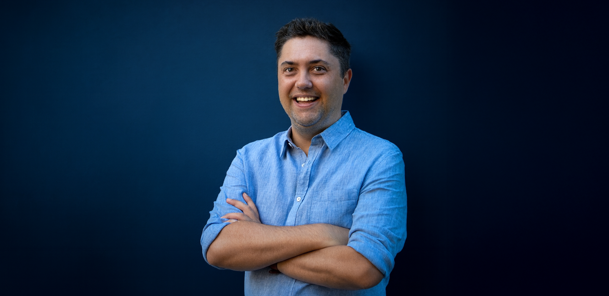 A young man with dark hair and a beard, smiling, wearing a light blue button-up shirt with sleeves rolled up, standing with arms crossed against a dark blue background.