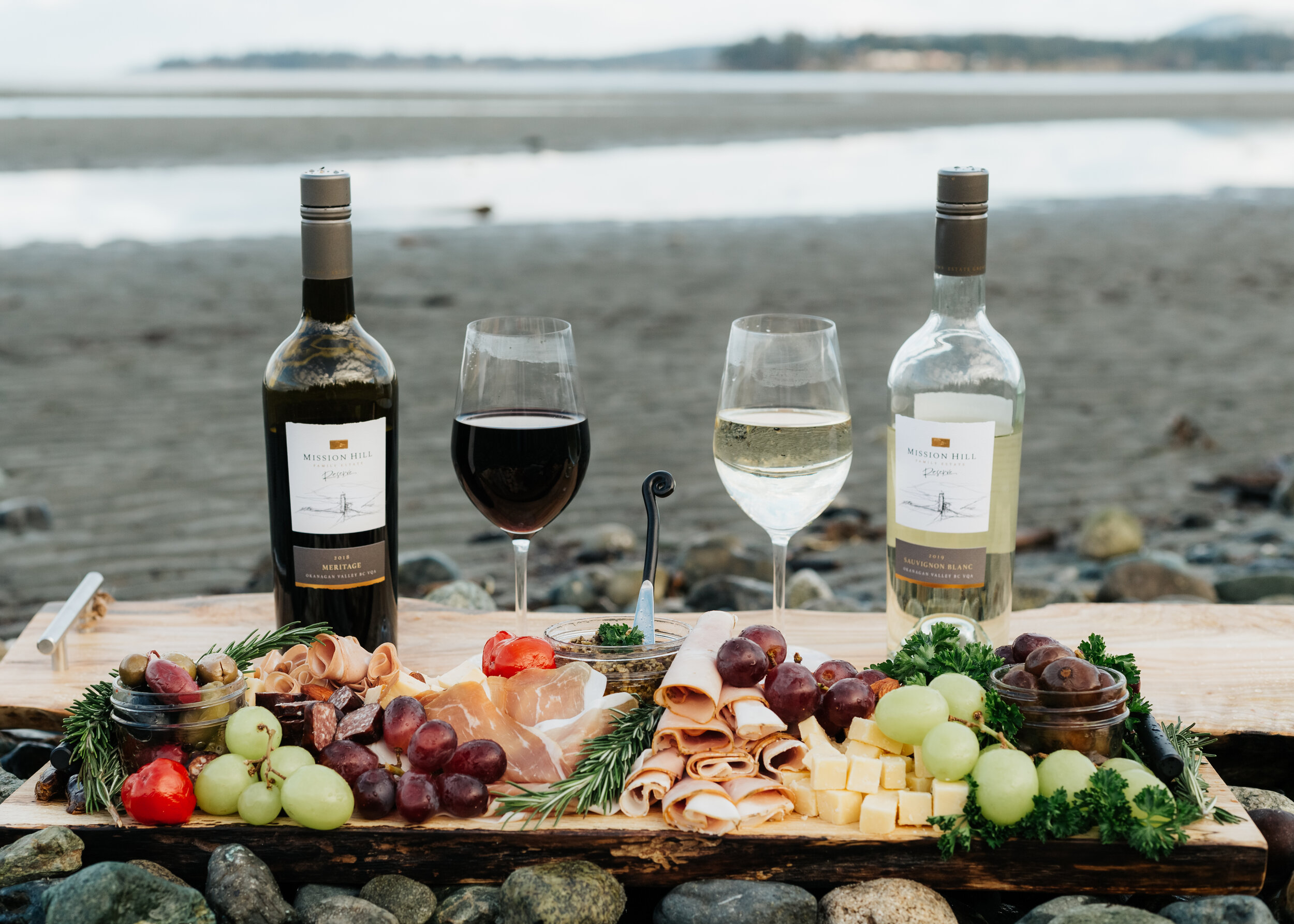 Outdoor picnic setting with two bottles of wine, glasses with red and white wine, charcuterie board of meats, cheeses, grapes, olives on a wooden plank by the beach.
