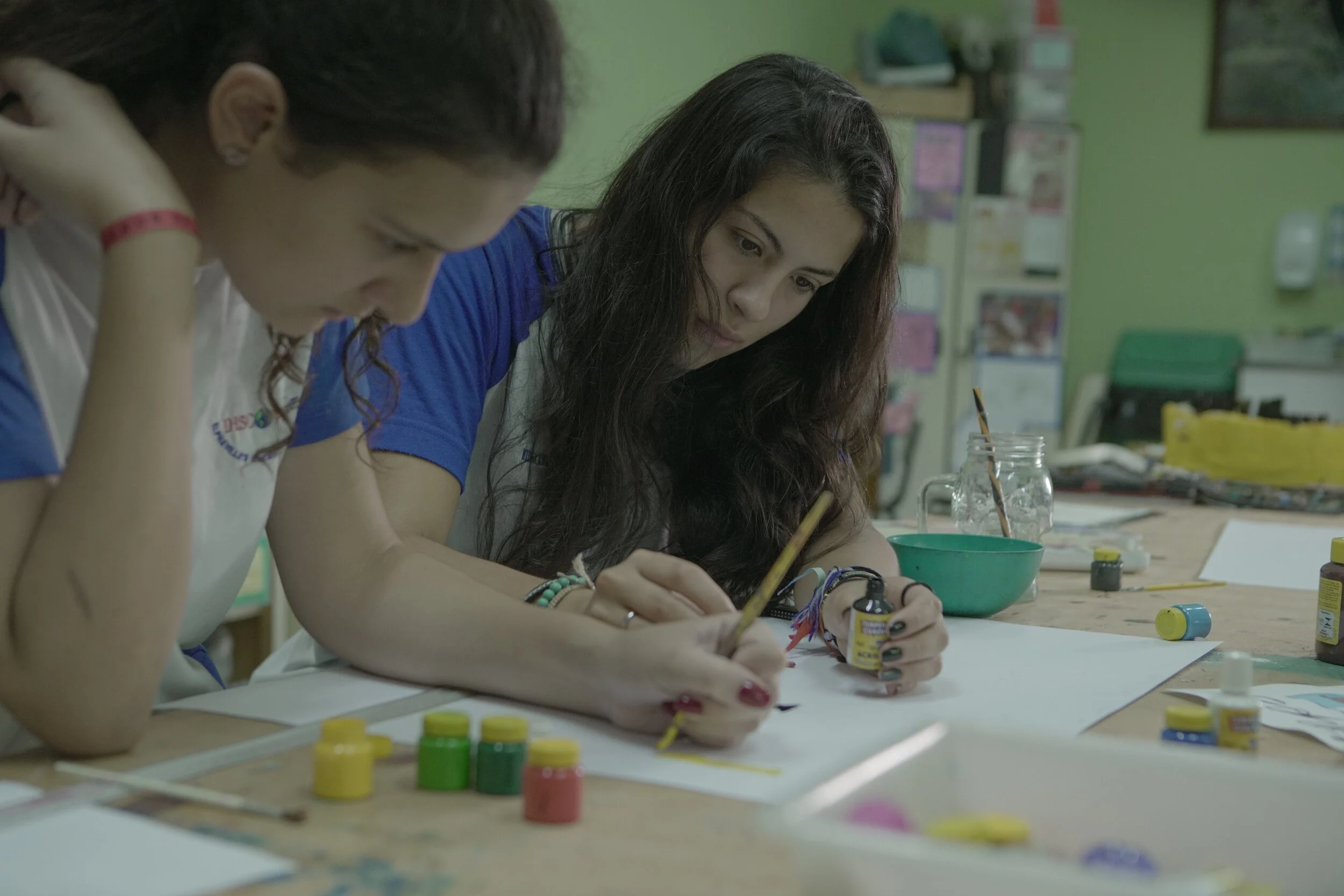 Two women working on a craft project at a table with paint jars and brushes in a classroom.