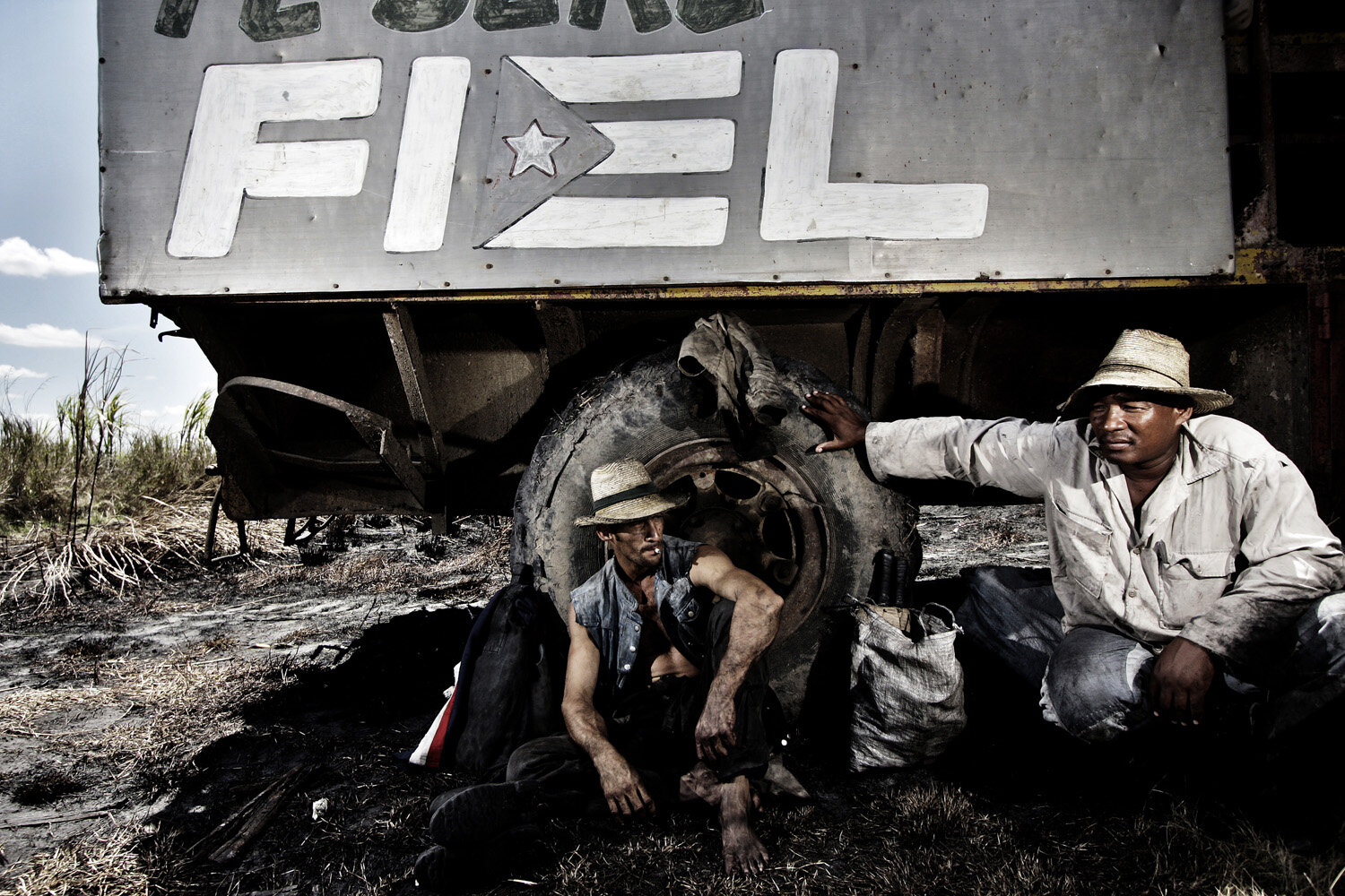  Maikel (L) and Juan Alberto (R) sugarcane cutter, sit under a truck where lunch is being served during a break. The sign on the truck reads, in Spanisch, both words: Fidel and Fiel, which means: Fidel and Faithfully. The two men have been working fo