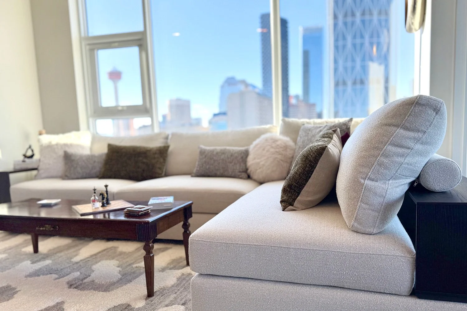 Light neutral furnishings, warm wood tones, and soft colourful accents create a comfortable seating area designed to showcase the city skyline views