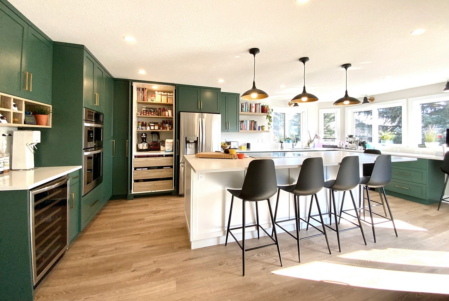 Modern kitchen with green cabinets, white island, black pendant lights, and wooden flooring, overlooking a bright windowed dining area.