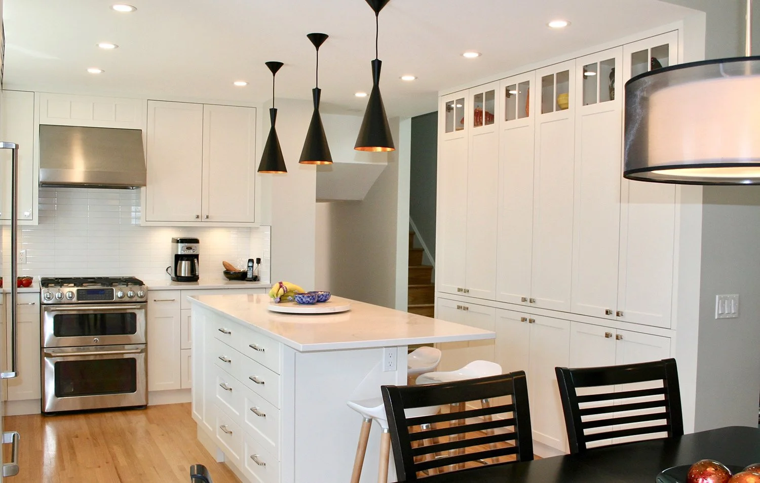 A modern white kitchen with stainless steel appliances and a shaker-style built-in pantry with glass display cabinets and interior lighting, accented with black cone pendant lights for contrast and style.