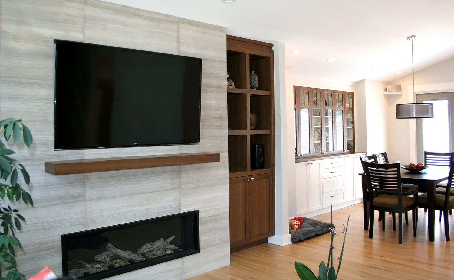 A limestone fireplace with an inset TV wall is framed by walnut display cabinets, while a built-in sideboard with illuminated glass cabinets provides elegant storage and display for china.