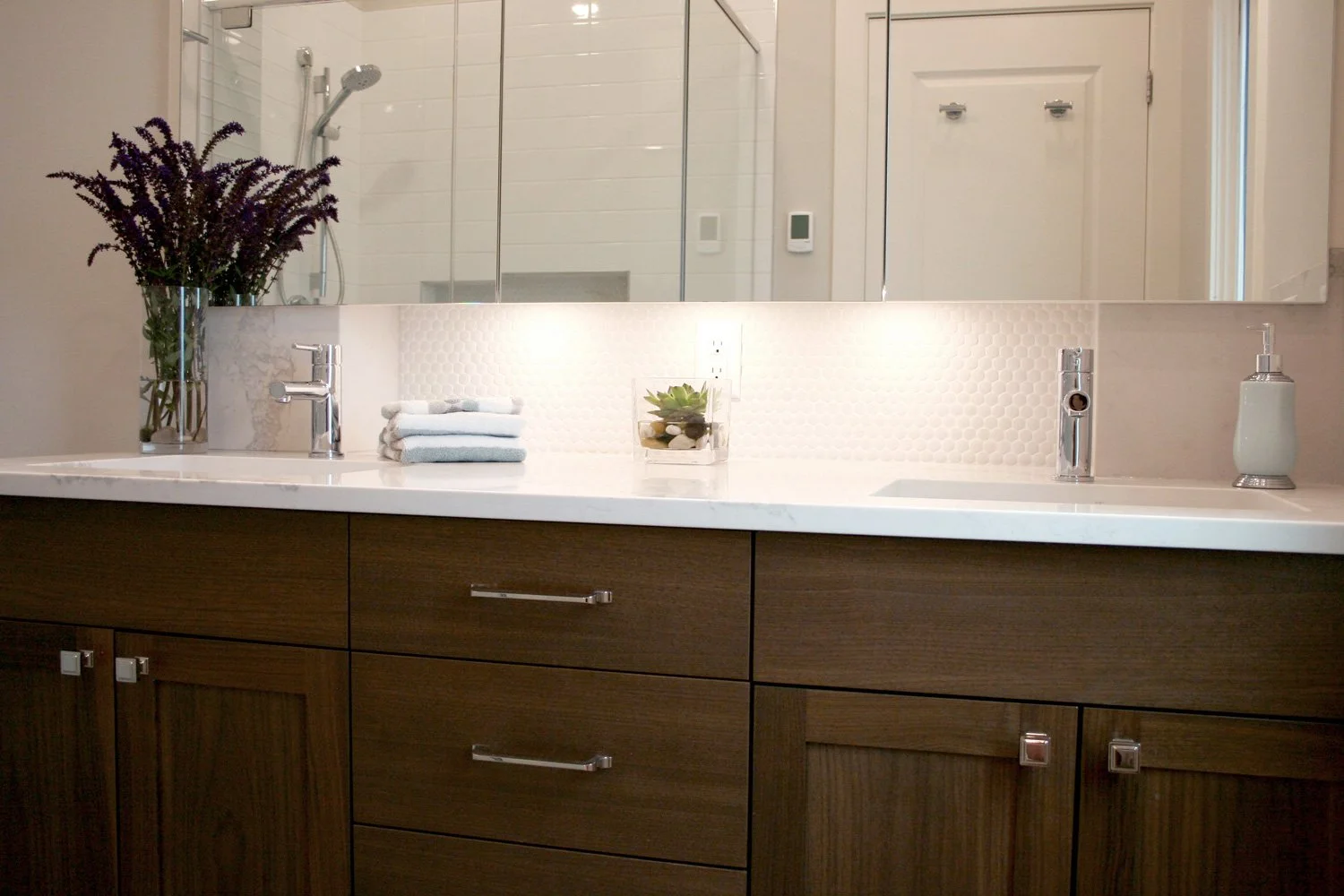 A modern bathroom design featuring a walnut vanity with a Calacatta quartz countertop, a large mirrored medicine cabinet, and under-cabinet lighting that illuminates the white penny round tile backsplash. The glass walk-in shower is tiled in large wh