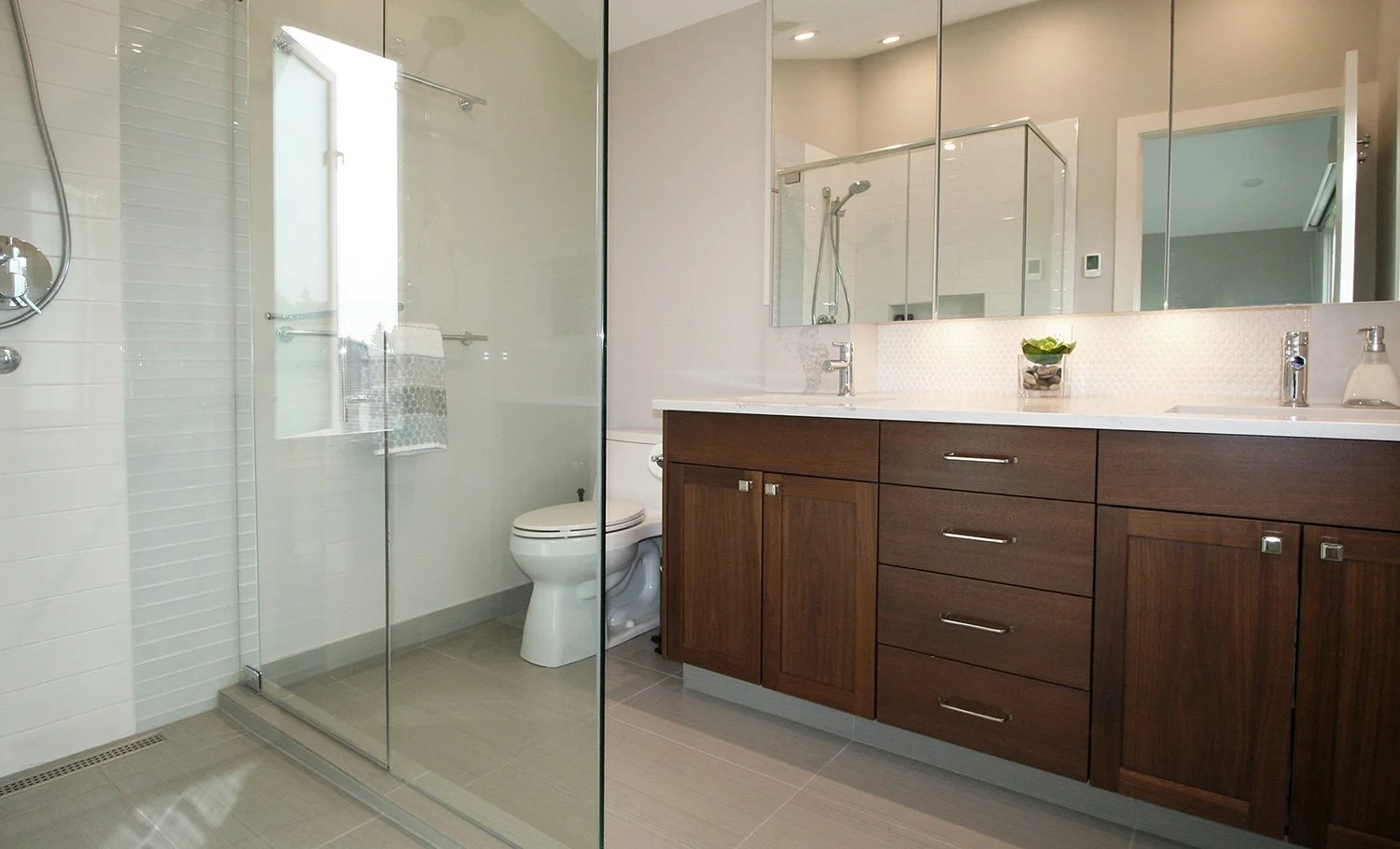 A modern bathroom design featuring a walnut vanity with a Calacatta quartz countertop, a large mirrored medicine cabinet, and under-cabinet lighting that illuminates the white penny round tile backsplash. The glass walk-in shower is tiled in large wh