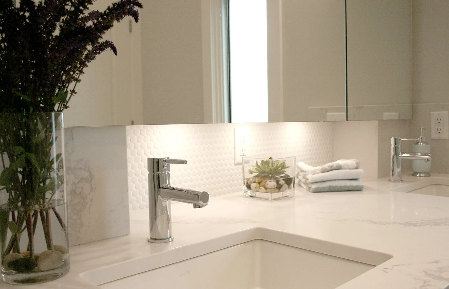 This modern bathroom vanity features a Calacatta quartz countertop, white penny round tile backsplash with under-cabinet lighting, and a large mirrored medicine cabinet, creating a bright spa-like bathroom with generous storage.