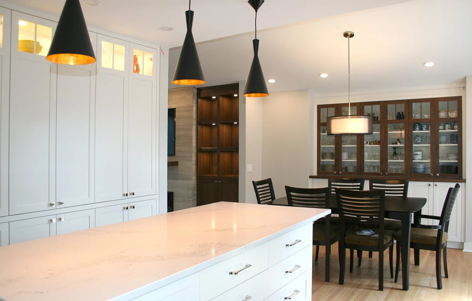 This modern transitional kitchen and dining space features white cabinetry and a built-in buffet with illuminated glass cabinets and dark wood accents. Black cone pendants and drum pendant lighting add contrast and visual interest.
