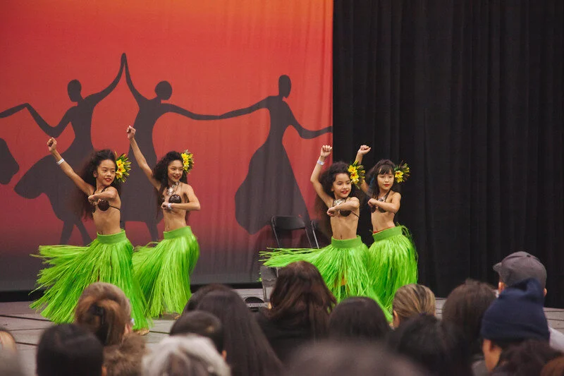 Four young Tahitian tamahine dancers in green more skirts and flower headpieces performing a dance on stage with a backdrop featuring silhouettes of dancers and a sunset gradient.