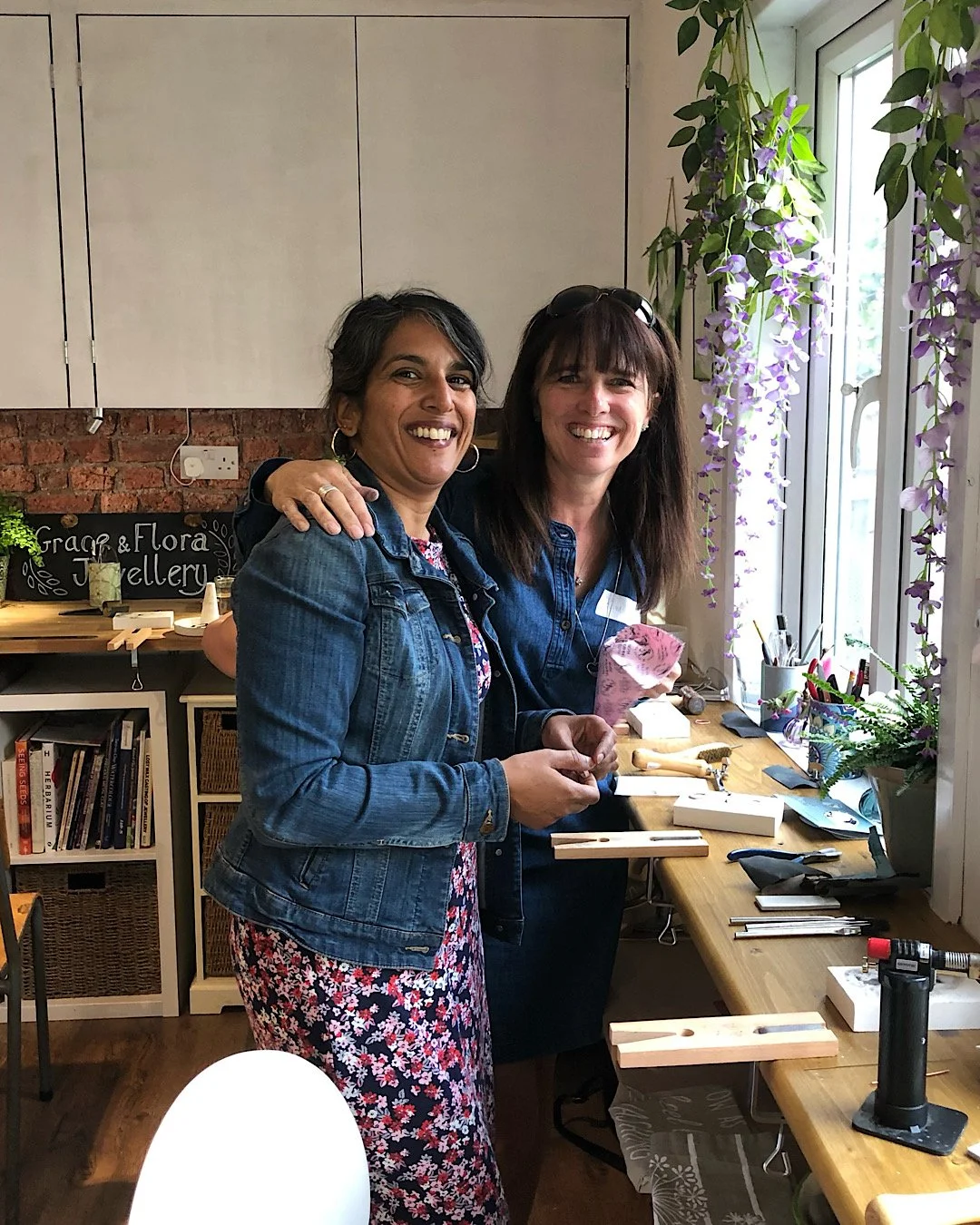 Two women smiling and standing close together in a nature inspired silver jewellery workshop, holding tools, with shelves, books, tools, and plants around them.