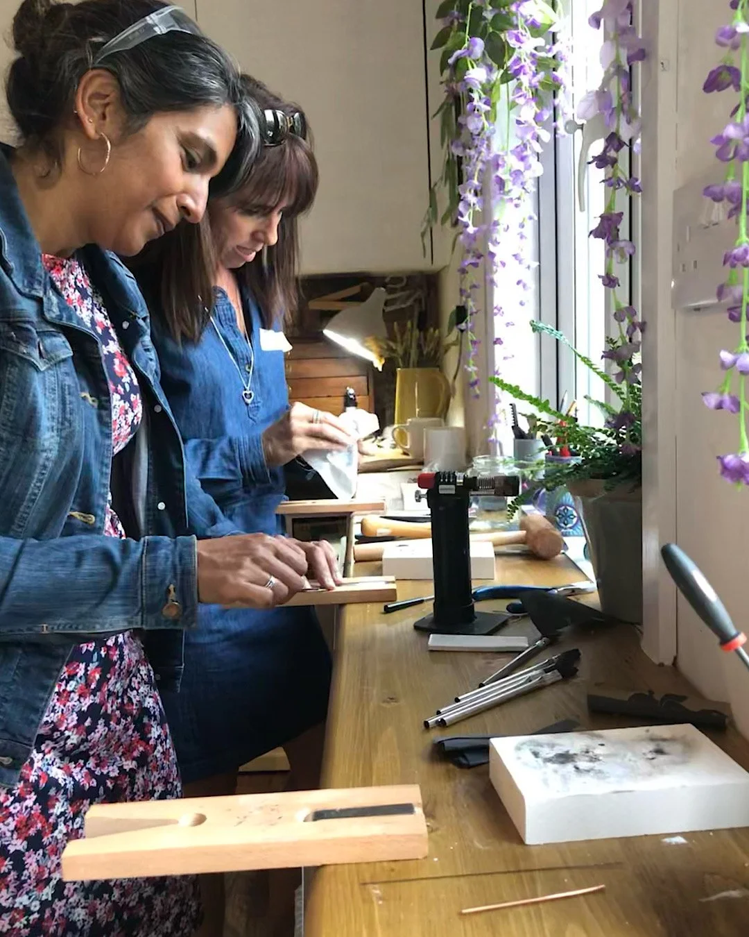 Two women working on creating silver stacking rings at a wooden table near a window with purple hanging flowers, tools, and supplies.