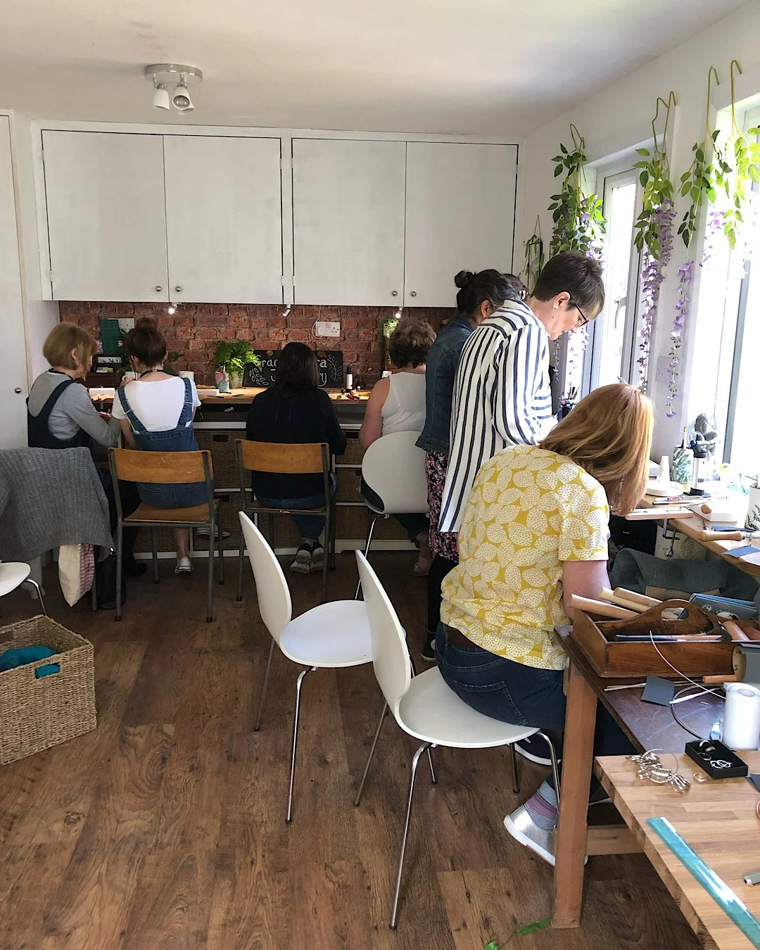 People gathered in a cozy room with natural light, making jewellery at tables, with plants and a brick wall in the background.