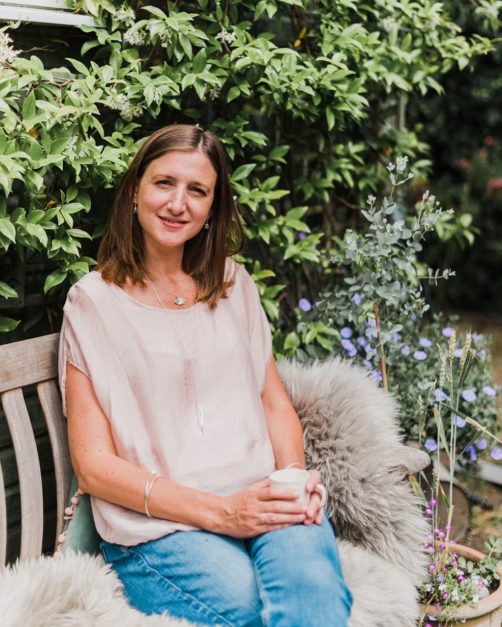handmade jewellery designer with silver nature necklaces sits on a bench in the sun with leaves behind outside the jewellery studio