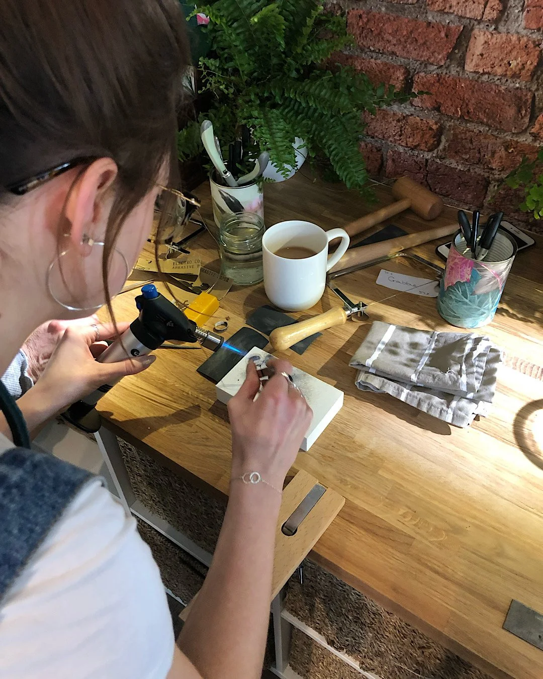 A woman on a silversmith jewellery making workshop and blow torch with wooden table with various tools, a cup of tea, and a plant in the background.