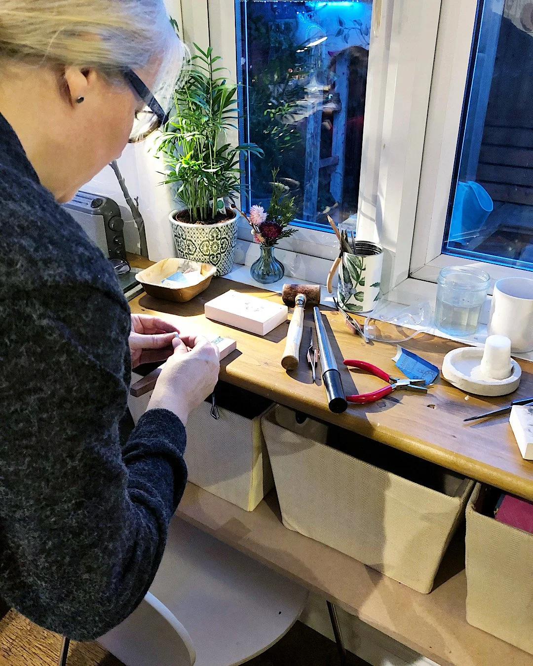 A person working at a wooden table with various near a window . The table has a potted plant, flowers, a container with pliers, and other silversmithing tools for jewellery making. The workspace is well-lit, and there are storage baskets below.