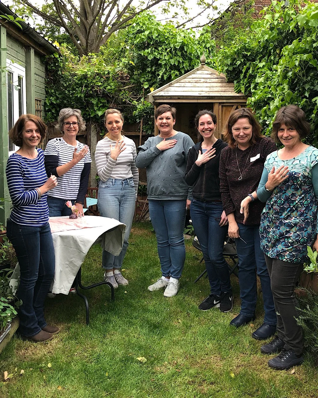 Group of eight women standing in a garden, with some placing their right hands over their hearts with their handmade stacking rings from the workshop, smiling, in front of green foliage and a small wooden shed.