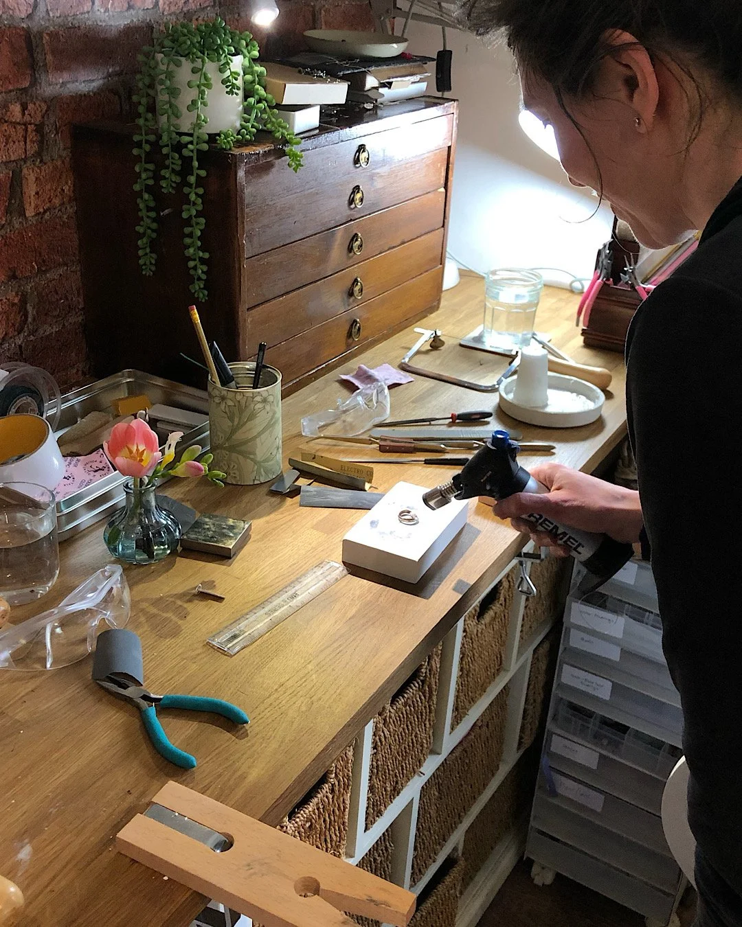 A person holding a soldering torch over a small jewellery piece at a cluttered wooden workspace with tools, glasses, and decorative items while working on stacking rings.