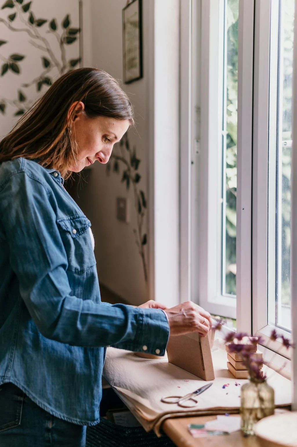 nature jewellery designer packaging jewellery in an eco-friendly box with recycled paper by a window and nature inspired studio - leaves are painted on the walls.