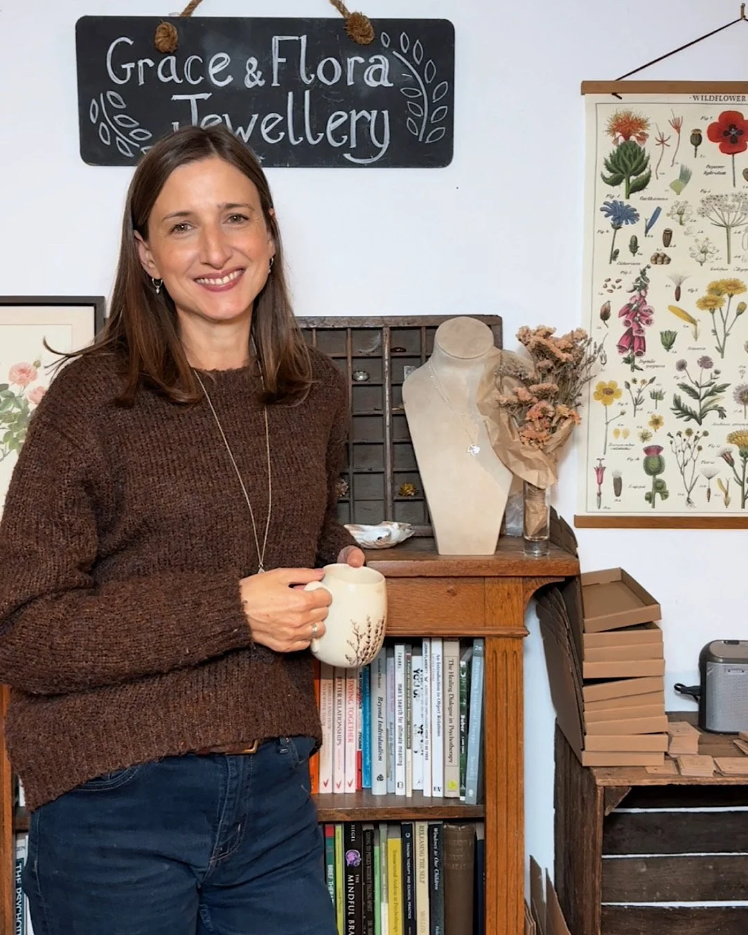Kate is a smiling woman with brown hair wearing a brown sweater and dark jeans, holding a white mug with grasses, standing in a cozy workshop, decorative plants, botanical print, and a chalkboard sign that reads 'Grace & Flora Jewellery'.