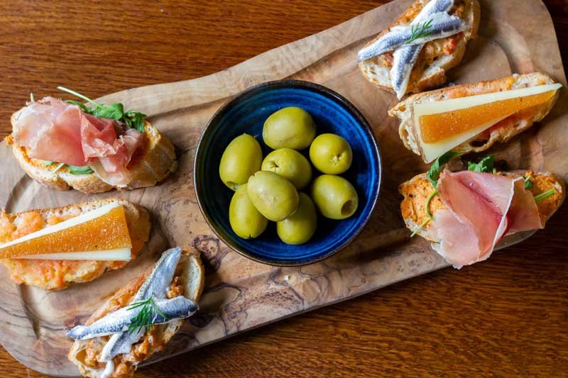  A platter of white anchovy fillets, Manchego cheese with Quince &amp; Serrano ham all served on Pan Catalán &amp; arranged around a small bowl of olives 
