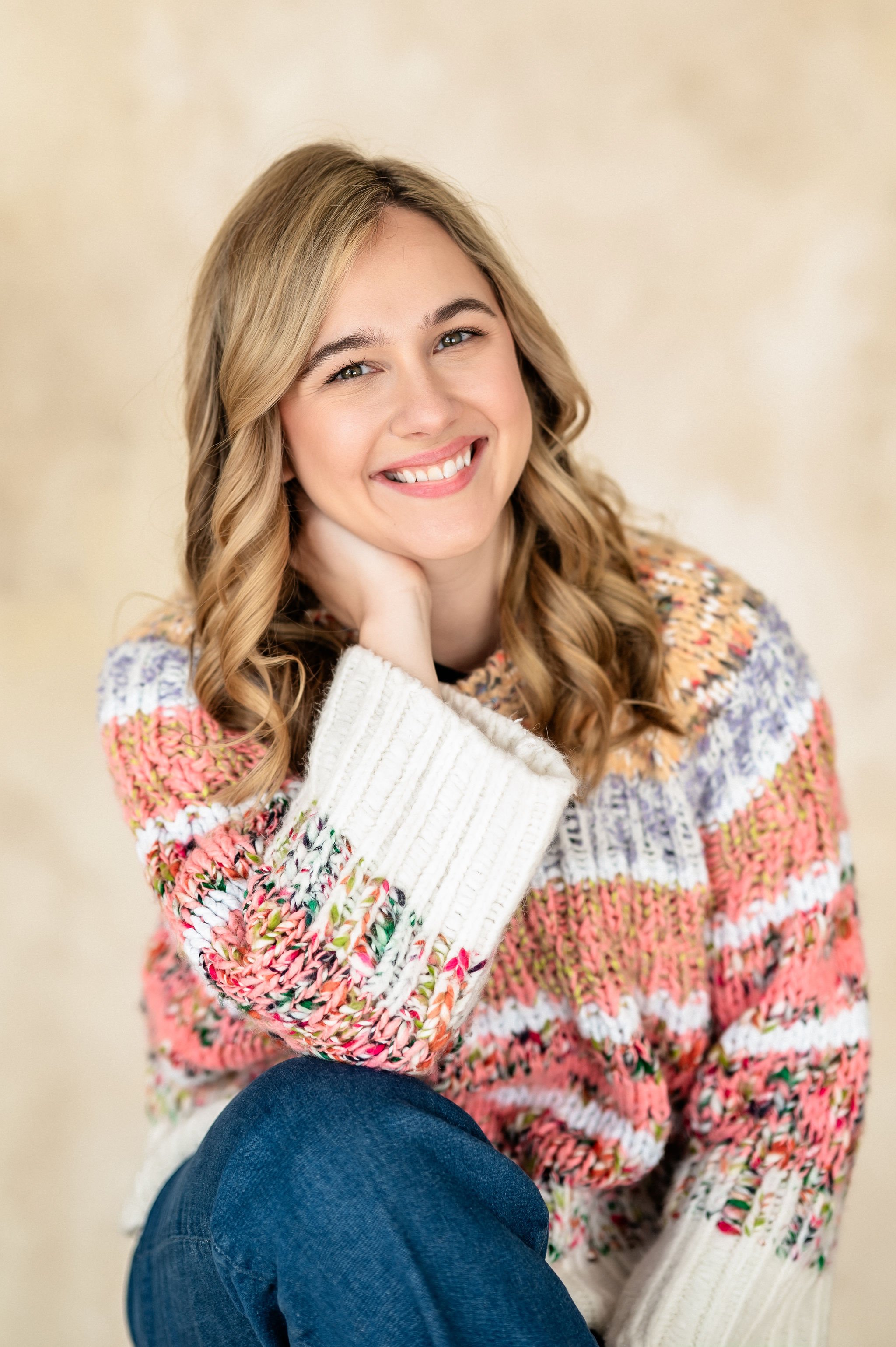 Young woman with blonde hair, wearing a colorful sweater and blue jeans, sitting and smiling at the camera.