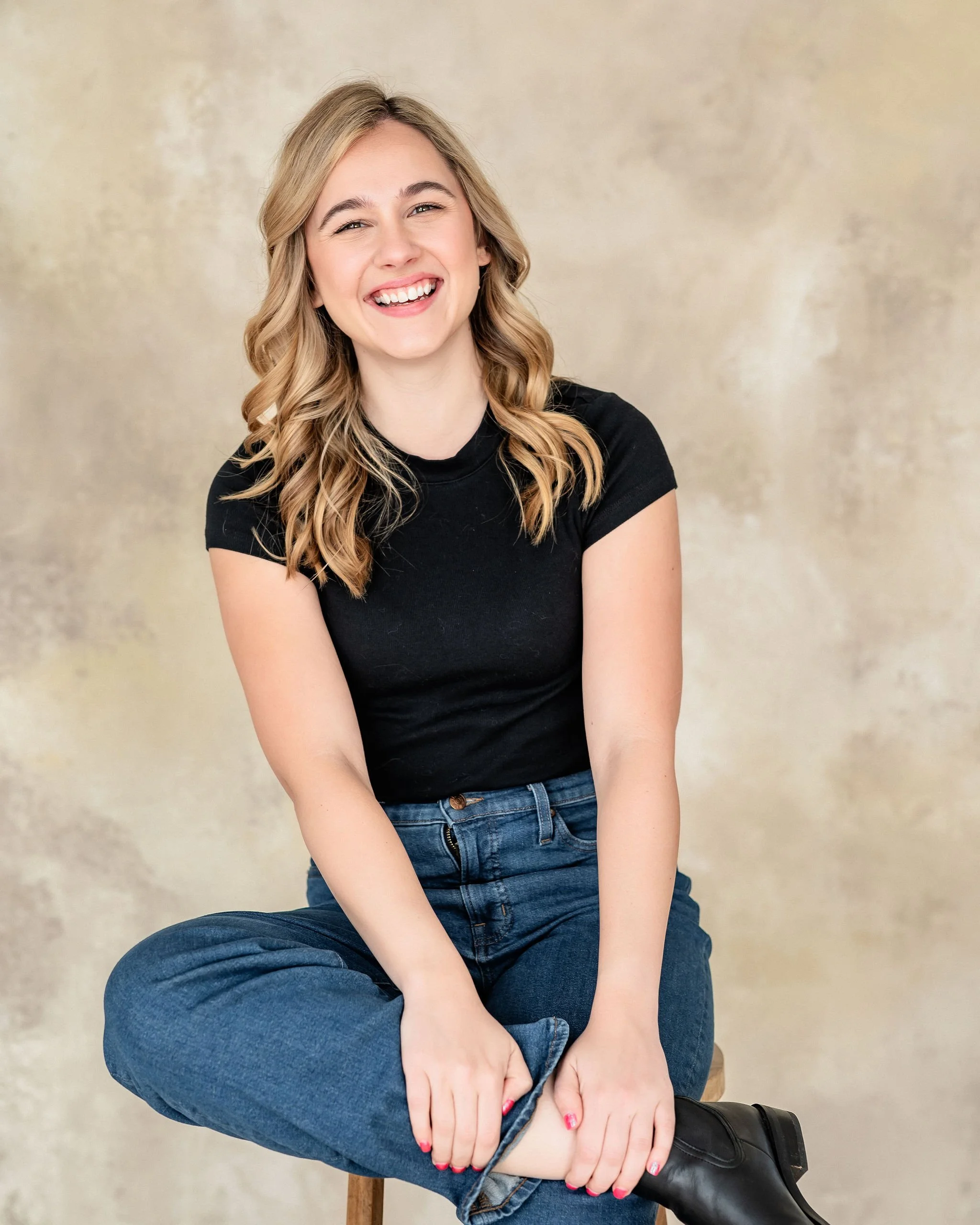 A young woman with blonde, wavy hair, sitting on a stool, smiling and wearing a black t-shirt, blue jeans, and black boots, against a beige background.
