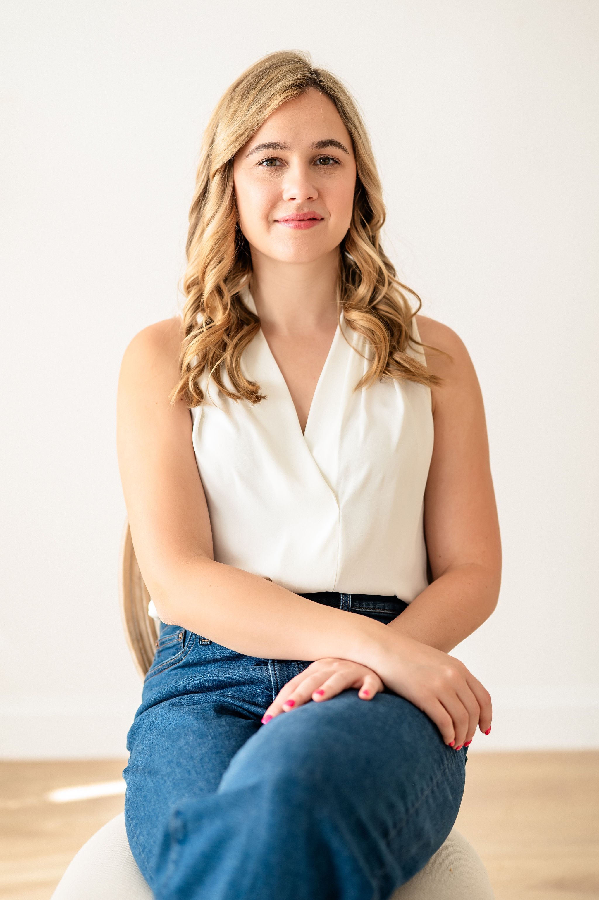 A young woman with blonde hair sitting on a white surface in front of a plain white wall, wearing a sleeveless cream-colored blouse and blue jeans, with her hands resting on her knee.