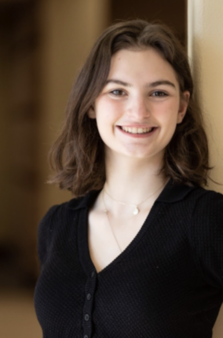 A young woman with shoulder-length brown hair smiling, wearing a black button-up top and a delicate necklace, standing indoors.